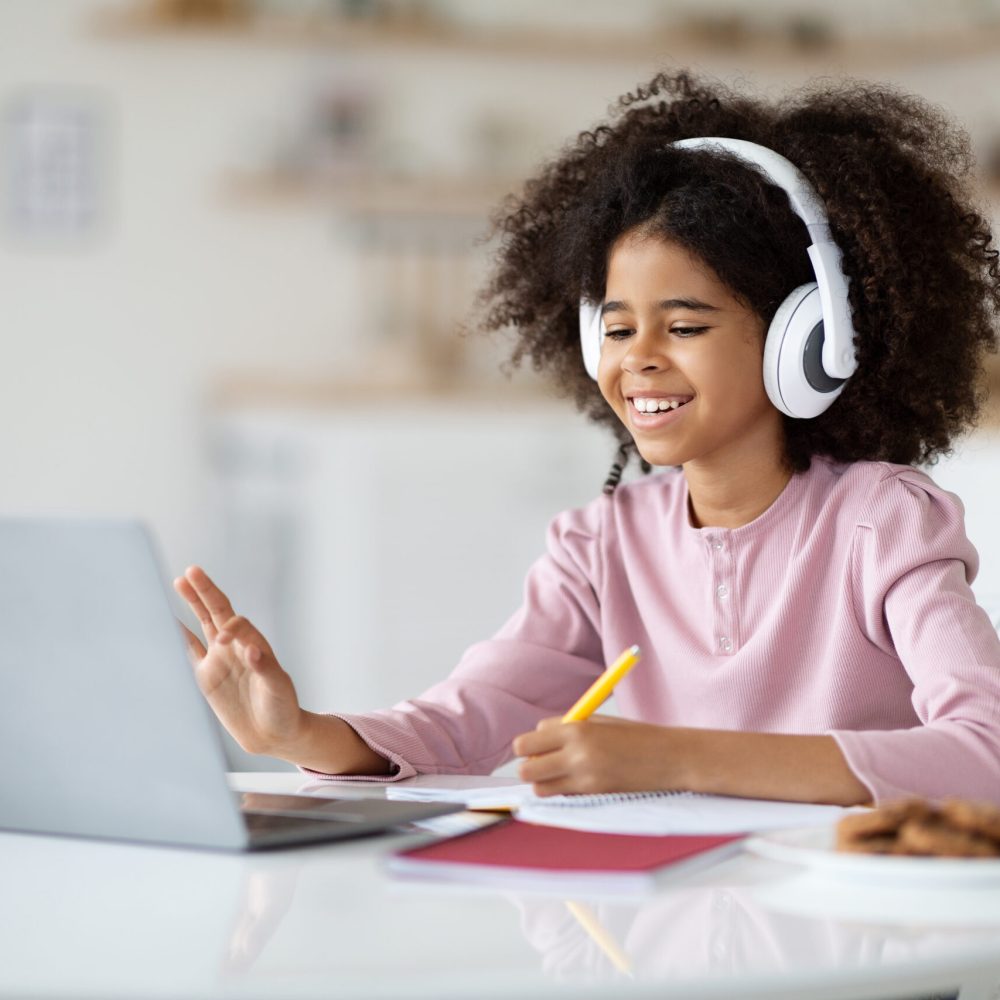 Positive black kid school girl with bushy hair having online party with friends while staying home, sitting at desk, drawing, using wireless headset, waving and smiling at computer screen, copy space